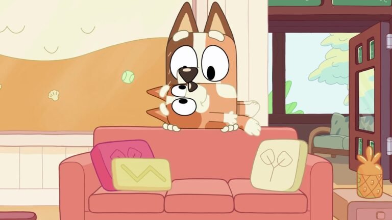 From the Bluey episode 'Keepy Uppy': Bluey and Bingo peek over the back of a pink couch with wide, focused eyes, mid-game, in their sunlit living room with pillows and a pineapple-shaped container nearby.